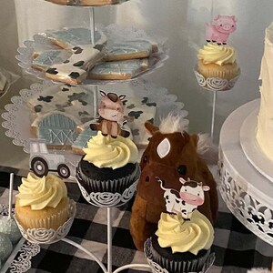 May include: A tiered cake stand with cupcakes decorated with farm animal toppers. The cupcakes have yellow icing and are topped with a brown horse, a pink pig, and a brown cow. The stand is decorated with white lace and is on a black and white checked tablecloth.