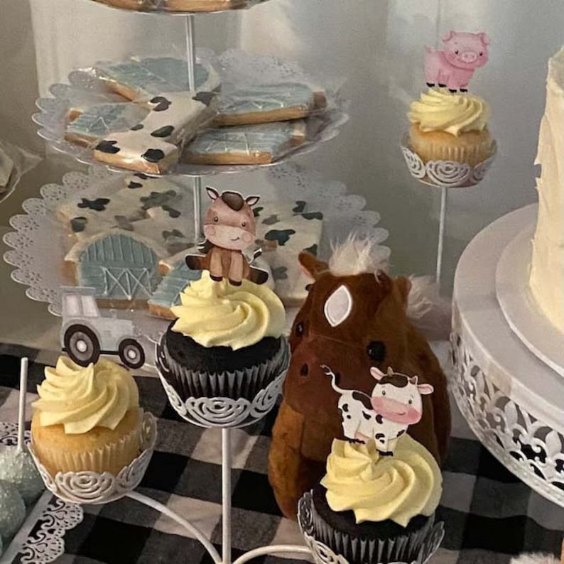 May include: A tiered cake stand with cupcakes decorated with farm animal toppers. The cupcakes have yellow icing and are topped with a brown horse, a pink pig, and a brown cow. The stand is decorated with white lace and is on a black and white checked tablecloth.