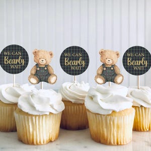 May include: Cupcakes with white frosting and decorative toppers. The toppers feature a teddy bear illustration and the text "We Can Bearly Wait" on a plaid background. The cupcakes are on a white surface.