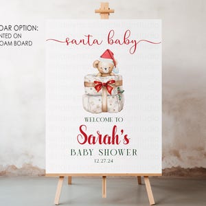 May include: A white rectangular sign on a wooden easel featuring the text santa baby, Sarah's Baby Shower, and the date 12.27.24. The design includes a watercolor illustration of a teddy bear wearing a red Santa hat sitting on two gift boxes.