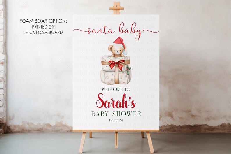 May include: A white rectangular sign on a wooden easel featuring the text santa baby, Sarah's Baby Shower, and the date 12.27.24. The design includes a watercolor illustration of a teddy bear wearing a red Santa hat sitting on two gift boxes.