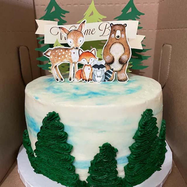 K&ouml;nnte beinhalten: Ein wei&szlig;er Kuchen, verziert mit gr&uuml;nen B&auml;umen und einem Topper mit einem Reh, einem Fuchs, einem Waschb&auml;ren und einem B&auml;ren. Der Topper sagt "Willkommen Baby".
