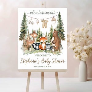 K&ouml;nnte beinhalten: Ein wei&szlig;es Willkommens-Schild mit Waldmotiv. Das Schild zeigt ein Reh, einen Fuchs, einen Waschb&auml;r, einen B&auml;ren und einen Baby-Strampler, der an einer W&auml;scheleine h&auml;ngt. Der Text auf dem Schild lautet "adventure awaits" und "Welcome to Stephanie's Baby Shower, November 9th, 2024".