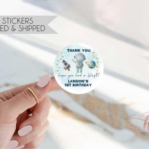 May include: Round thank you sticker with a blue astronaut, rocket, and stars on a white background. The text reads "Thank you, hope you had a blast! Landon's 1st Birthday".