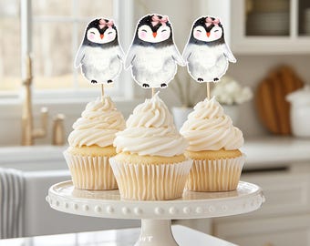 May include: Three cupcakes with white frosting are displayed on a white cake stand. Each cupcake is topped with a penguin-shaped decoration. The penguins are black and white with pink bows.