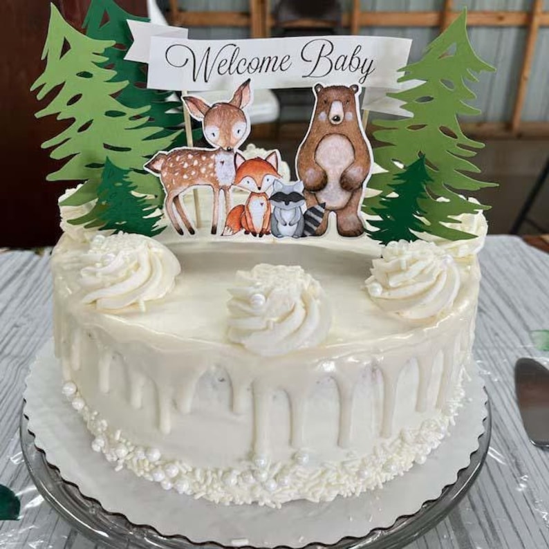 K&ouml;nnte beinhalten: Ein wei&szlig;er Kuchen mit wei&szlig;em Frosting und einem wei&szlig;en Drip. Der Kuchen ist mit einem Waldtier-Motiv verziert, darunter ein Reh, ein Fuchs, ein Waschb&auml;r und ein B&auml;r. Der Kuchen hat auch ein Banner mit der Aufschrift "Willkommen Baby".