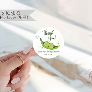 May include: A 2-inch round sticker with a baby shower theme. The sticker features a green pea pod with two smiling peas, the text "Thank You!", and "Sabrina's baby shower 03/24/24". The sticker is white with green and pink accents.