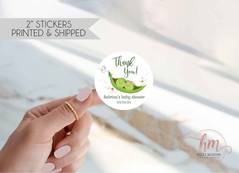 May include: A 2-inch round sticker with a baby shower theme. The sticker features a green pea pod with two smiling peas, the text "Thank You!", and "Sabrina's baby shower 03/24/24". The sticker is white with green and pink accents.