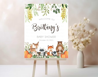 May include: A baby shower welcome sign on a wooden easel. The sign features the text "Welcome to Brittany's Baby Shower October 10, 2021" with watercolor illustrations of woodland animals and pumpkins. Green foliage decorates the top.