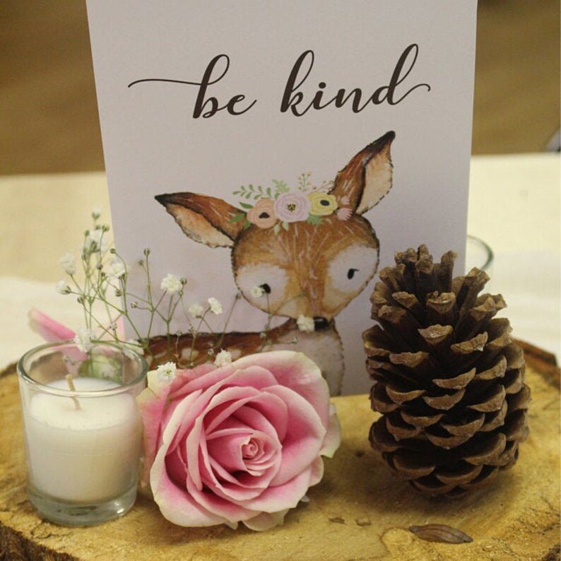 Puede incluir: Un centro de mesa de madera con una rosa rosa, una vela blanca peque&ntilde;a, una pi&ntilde;a y una tarjeta con una ilustraci&oacute;n de un ciervo y el texto "be kind".