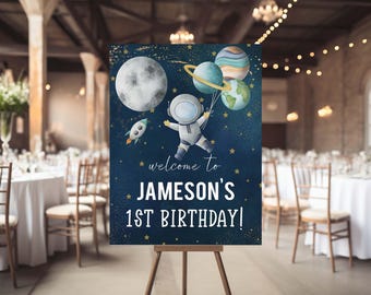 May include: A blue and gold welcome sign with a cartoon astronaut holding balloons shaped like planets. The sign says "Welcome to Jameson's 1st Birthday!"