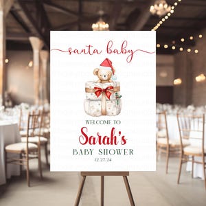 May include: A white rectangular sign featuring the red text santa baby at the top, a watercolor illustration of a teddy bear wearing a red Santa hat sitting on two wrapped gifts, and the text WELCOME TO Sarah's BABY SHOWER 12.27.24 below.