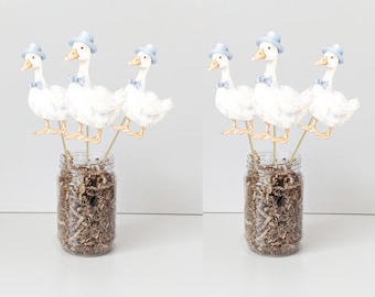 May include: A pair of glass jars filled with brown shredded paper, each holding four white duck figurines. The ducks wear blue hats and bow ties. The ducks are attached to thin gold-colored sticks.