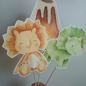 May include: Three watercolor dinosaur cupcake toppers. A green triceratops, a yellow triceratops, and a brown volcano.
