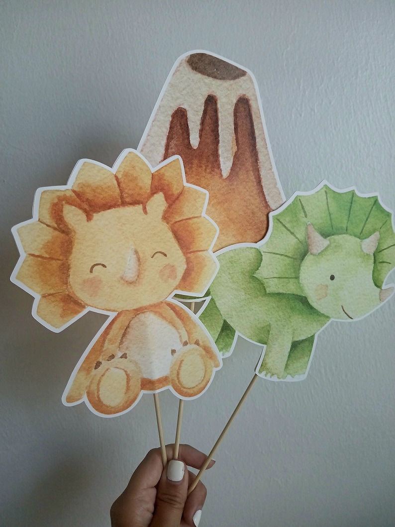 May include: Three watercolor dinosaur cupcake toppers. A green triceratops, a yellow triceratops, and a brown volcano.