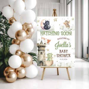 May include: A baby shower sign on a wooden easel, featuring a watercolor illustration of dragons, clouds, and stars. The sign reads "Hatching Soon! Welcome to Joella's Baby Shower 01.31.2026". White and gold balloons are arranged to the left.