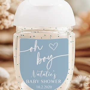 Puede incluir: Un frasco transparente de desinfectante de manos con tapa blanca. Una etiqueta azul dice "oh boy" con un coraz&oacute;n, "Natalie's BABY SHOWER" y la fecha "10.2.2020". La botella tiene peque&ntilde;as motas azules dispersas.