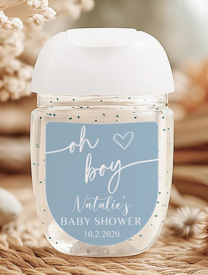 Puede incluir: Un frasco transparente de desinfectante de manos con tapa blanca. Una etiqueta azul dice "oh boy" con un coraz&oacute;n, "Natalie's BABY SHOWER" y la fecha "10.2.2020". La botella tiene peque&ntilde;as motas azules dispersas.