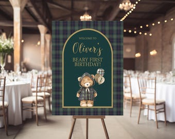 May include: A welcome sign for a first birthday party. The sign features a teddy bear wearing a plaid jacket, with the text "Welcome to Oliver's Beary First Birthday!" The sign is framed with a green and navy plaid pattern.