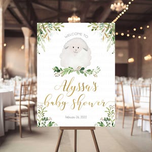 May include: A white welcome sign with green foliage and a white lamb illustration. The sign reads "Welcome to Alyssa's Baby Shower, February 26, 2022".