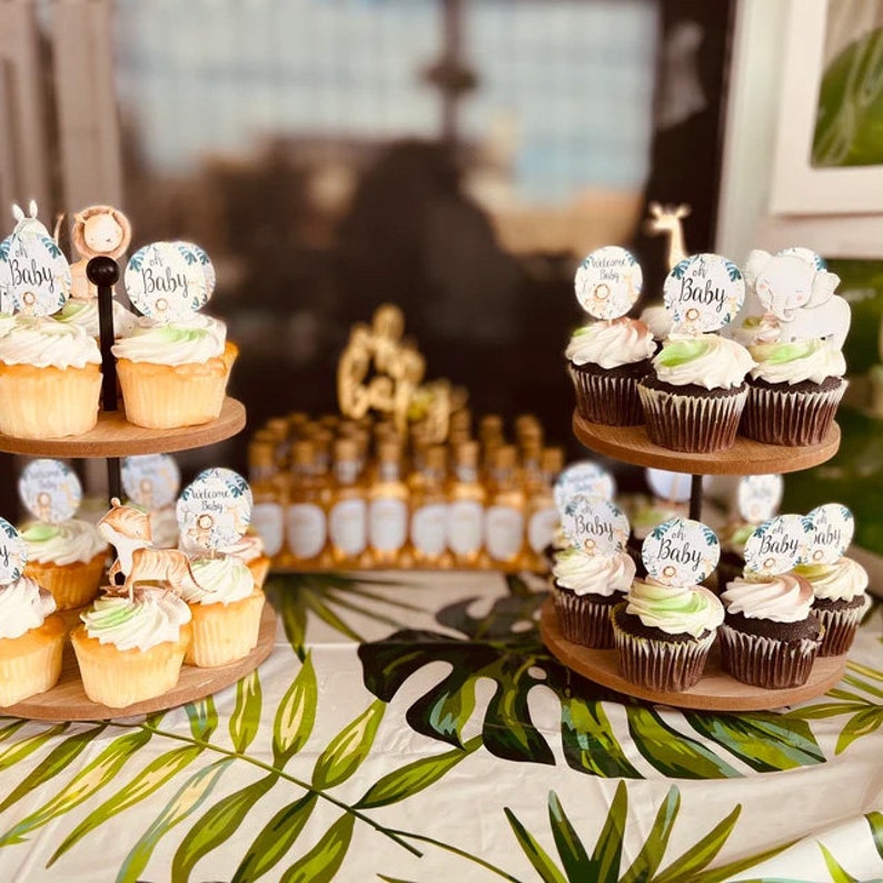 May include: Two-tiered cake stand with cupcakes decorated with jungle animal themed toppers. The cupcakes are frosted with white and green frosting. The stand is on a table with a green and white tropical leaf pattern tablecloth. The cupcakes have toppers that say "Welcome Baby" and "Oh Baby".
