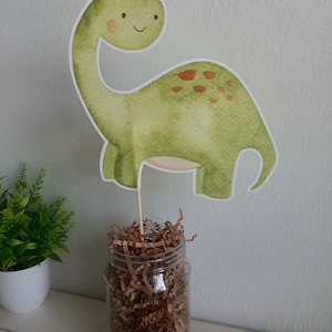 May include: A green dinosaur party centerpiece with brown spots, a smiling face, and a long neck. The dinosaur is made of paper and is attached to a wooden stick. The stick is inserted into a clear glass jar filled with brown shredded paper.