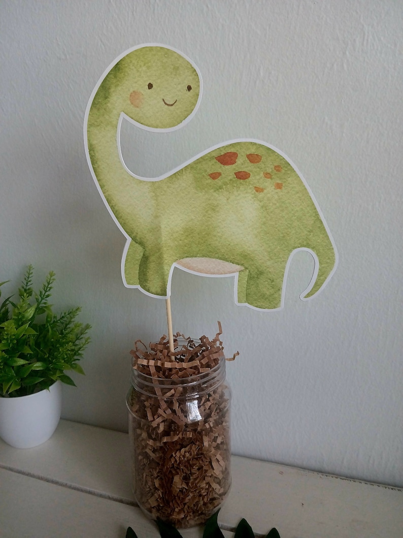 May include: A green dinosaur party centerpiece with brown spots, a smiling face, and a long neck. The dinosaur is made of paper and is attached to a wooden stick. The stick is inserted into a clear glass jar filled with brown shredded paper.