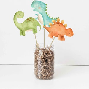 May include: Three watercolor dinosaur figurines on wooden sticks in a clear glass jar filled with brown shredded paper. The dinosaurs are green, blue, and orange.