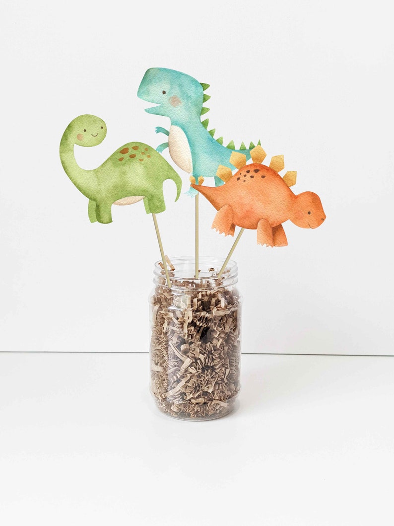 May include: Three watercolor dinosaur figurines on wooden sticks in a clear glass jar filled with brown shredded paper. The dinosaurs are green, blue, and orange.
