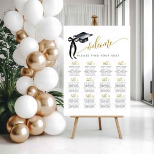 May include: A graduation welcome sign with a black mortarboard and ribbon graphic, gold script, and guest seating chart. The sign is displayed on a wooden easel, with white and gold balloons in the background.