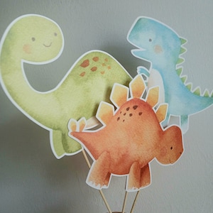May include: Three watercolor dinosaur cutouts on wooden sticks. The dinosaurs are a green brachiosaurus, an orange stegosaurus, and a blue velociraptor.