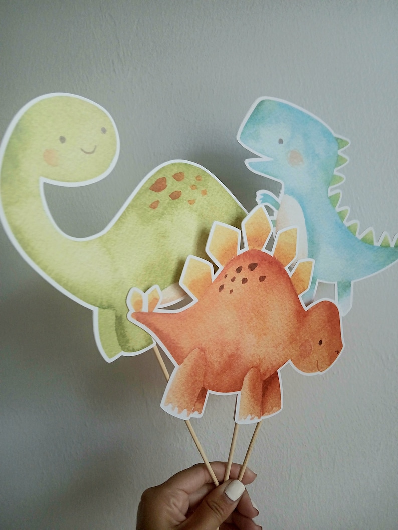 May include: Three watercolor dinosaur cutouts on wooden sticks. The dinosaurs are a green brachiosaurus, an orange stegosaurus, and a blue velociraptor.