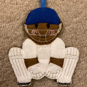 May include: A felt cricket player decoration. The ornament features a blue helmet, brown face, and white protective gear. The ornament has a blue ribbon for hanging. The cricket player is in a crouched position.