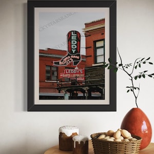 May include: A framed photograph of a vintage storefront with a neon sign shaped like a boot. The sign reads "Leddy's Hand Made Boots" and "Leddy's Boots & Saddlery".