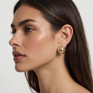 May include: A polished gold-tone spherical stud earring with a smooth, reflective surface. The earring features a rounded, ball-shaped design that sits flush against the earlobe, providing a simple and classic metallic accessory for daily wear.