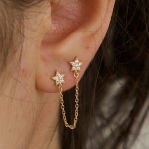 May include: Gold-colored star earrings with a chain connecting two star-shaped studs. The stars are embellished with small, clear gemstones. The earrings are shown on an ear.