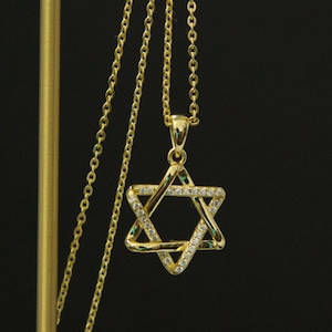 May include: A gold-colored necklace with a Star of David pendant. The pendant is encrusted with small, clear crystals and green accents. The necklace hangs against a dark background.