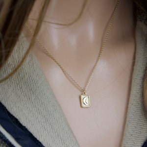 May include: A gold-colored necklace with a rectangular pendant featuring a crescent moon and sunburst design. The necklace is worn on a person with long, light brown hair and a tweed jacket with a navy blue trim.