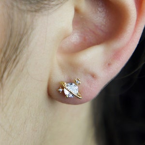 May include: Gold-toned stud earrings with a clear crystal in the shape of a planet with a ring around it. The earrings are set against a dark background.