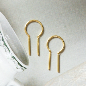 May include: A pair of gold-colored earrings. Each earring features a curved top with a straight bar pendant. The earrings are displayed on a light-colored surface, with a teacup and lace in the background. The earrings have a minimalist design.