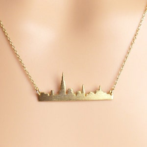 May include: A gold necklace featuring a delicate chain and a pendant shaped like a city skyline.