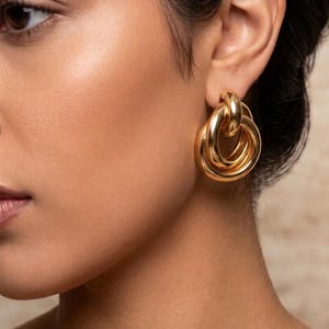 May include: A gold-toned earring featuring a design of three interlocking circular hoops of varying sizes. The earring has a polished, reflective metallic finish and is secured to the earlobe with a post-style backing, creating a layered appearance.