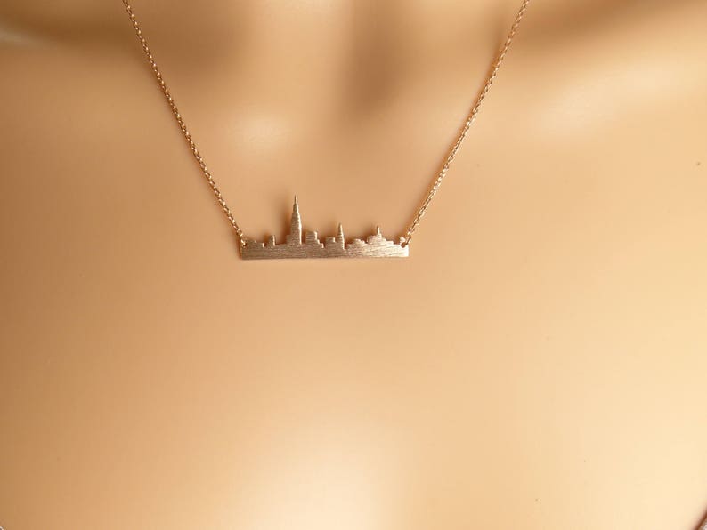 Puede incluir: Un collar de oro con un delicado colgante de ciudad. El colgante es una silueta de un horizonte urbano con edificios altos.