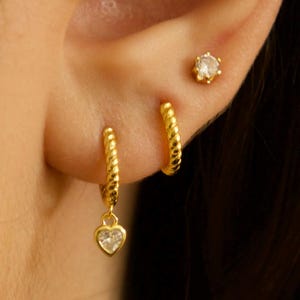 May include: Gold hoop earrings with a small heart charm dangling from one earring. The heart charm has a clear crystal in the center. The earrings are shown in a person's ear.