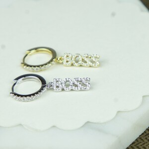 May include: A pair of earrings with a hoop and a dangling charm that says "BOSS" in sparkly letters. One earring is gold and the other is silver.