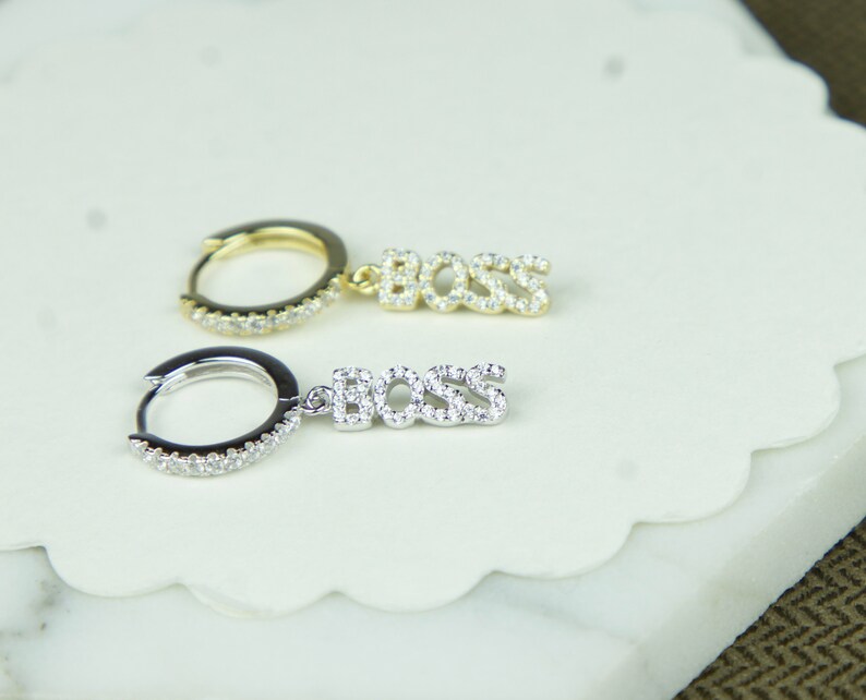 May include: A pair of earrings with a hoop and a dangling charm that says "BOSS" in sparkly letters. One earring is gold and the other is silver.
