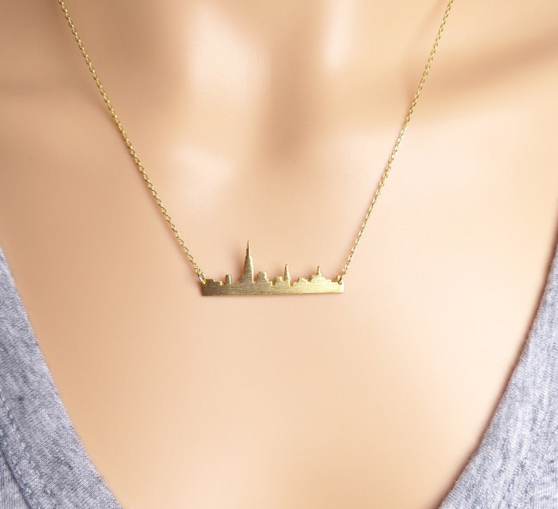 Puede incluir: Un collar de oro con un colgante de ciudad. El colgante es una silueta de un horizonte urbano.