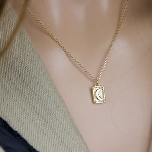 May include: Gold-colored necklace with a rectangular pendant featuring a crescent moon design. The pendant is framed by a sunburst pattern. The necklace is worn against a neutral-toned background.