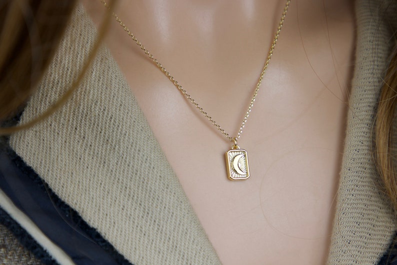 May include: Gold-colored necklace with a rectangular pendant featuring a crescent moon design. The pendant is framed by a sunburst pattern. The necklace is worn against a neutral-toned background.