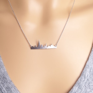 Puede incluir: Un collar de plata con un colgante de ciudad. El colgante es una silueta detallada de un horizonte urbano.